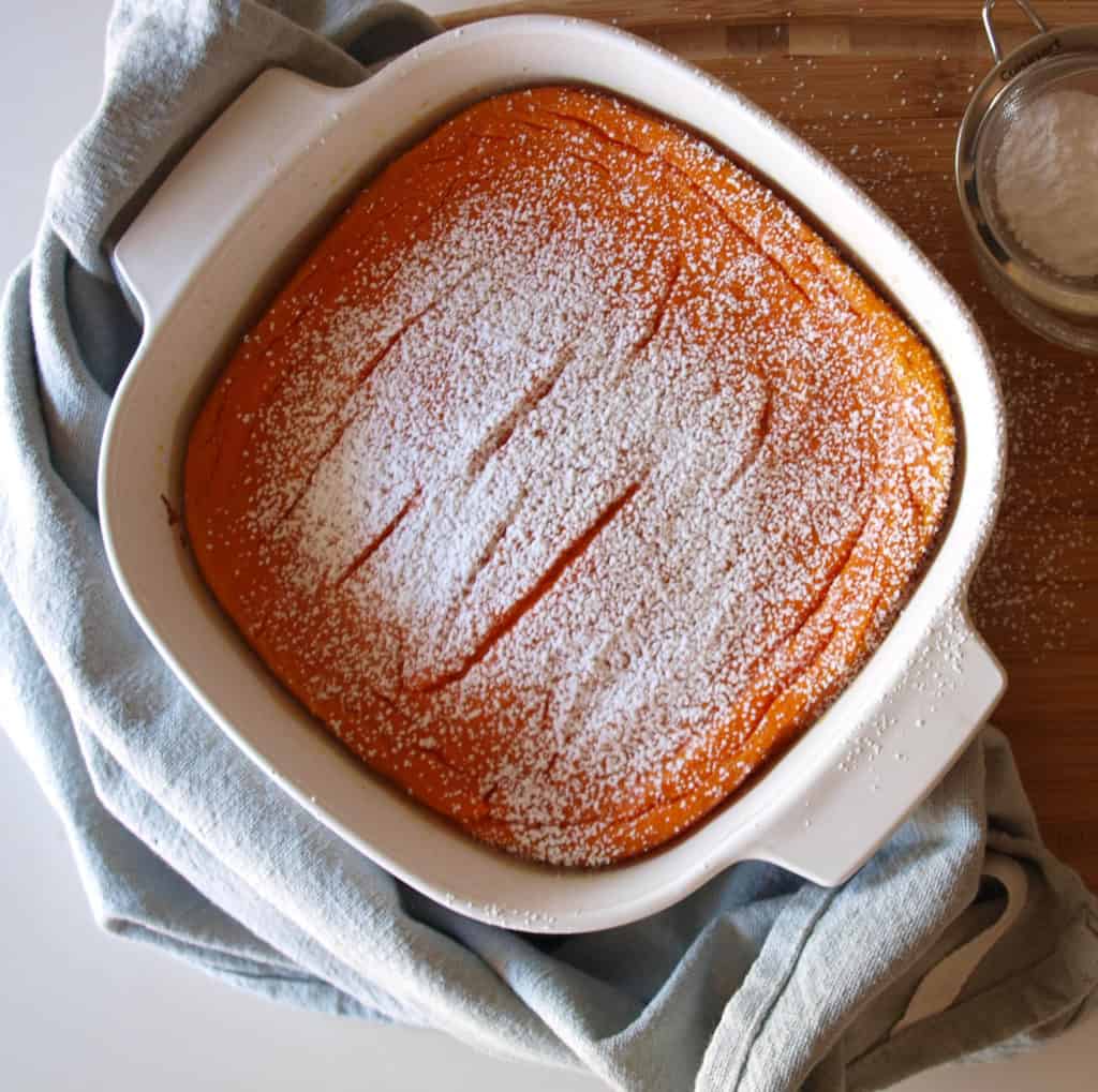 An easter dinner side Carrot Souffle Topped With Powdered Sugar on top of a wooden cutting board. A blue dish towel and powdered sugar sits next to the souffle.