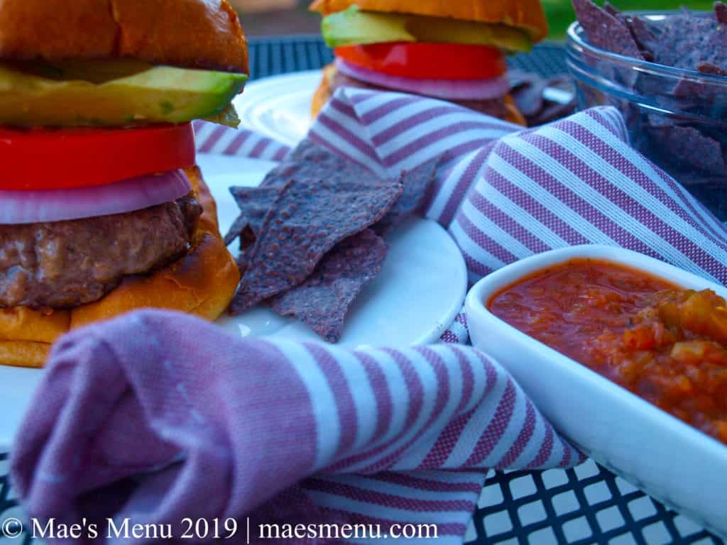 Juicy hamburgers sitting on white plates next to a purple dish towel and bowl of blue corn tortilla chips.