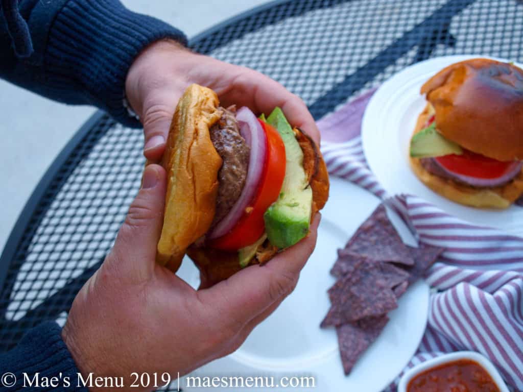 Male hands holding a burger over a table with a white plate, blue corn tortilla chips, salsa, a purple striped dish towel, and another burger on it. 