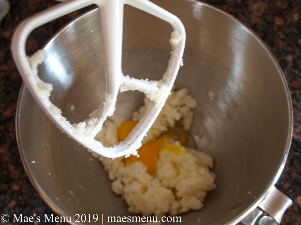 A white stand mixer paddle attachment over a silver mixing bowl with creamed sugar and an egg in it. 