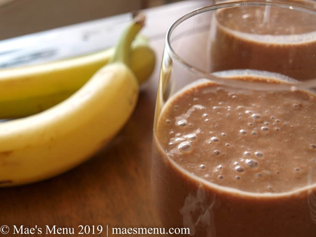 Two glasses of healthy chocolate smoothie with banana and coconut next to 2 bananas.