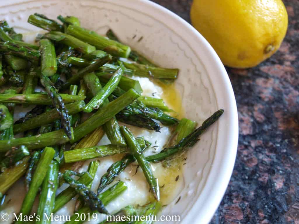White porcelain bowl of white grits with ricotta make a perfect easter dinner side.. Roasted asparagus with a lemon drizzle is on top of the grits. A lemon sits to the back right of bowl.