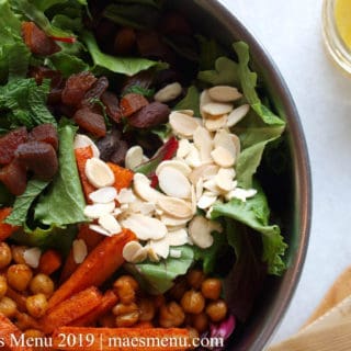 Metal bowl of salad with slivered almonds, roasted chickpeas & carrots, dried apricots, and mint with salad tongs and lemon dressing on the white granite counter next to the bowl.