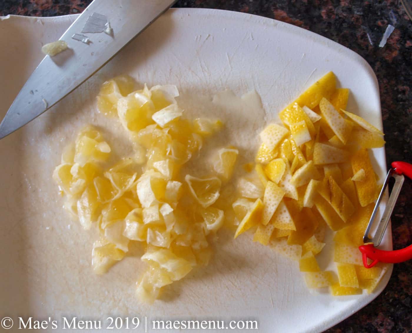 Chopped peeled lemons and chopped lemon zest on a white cutting board. Chefs knife at the top and red vegetable peeler on the right.