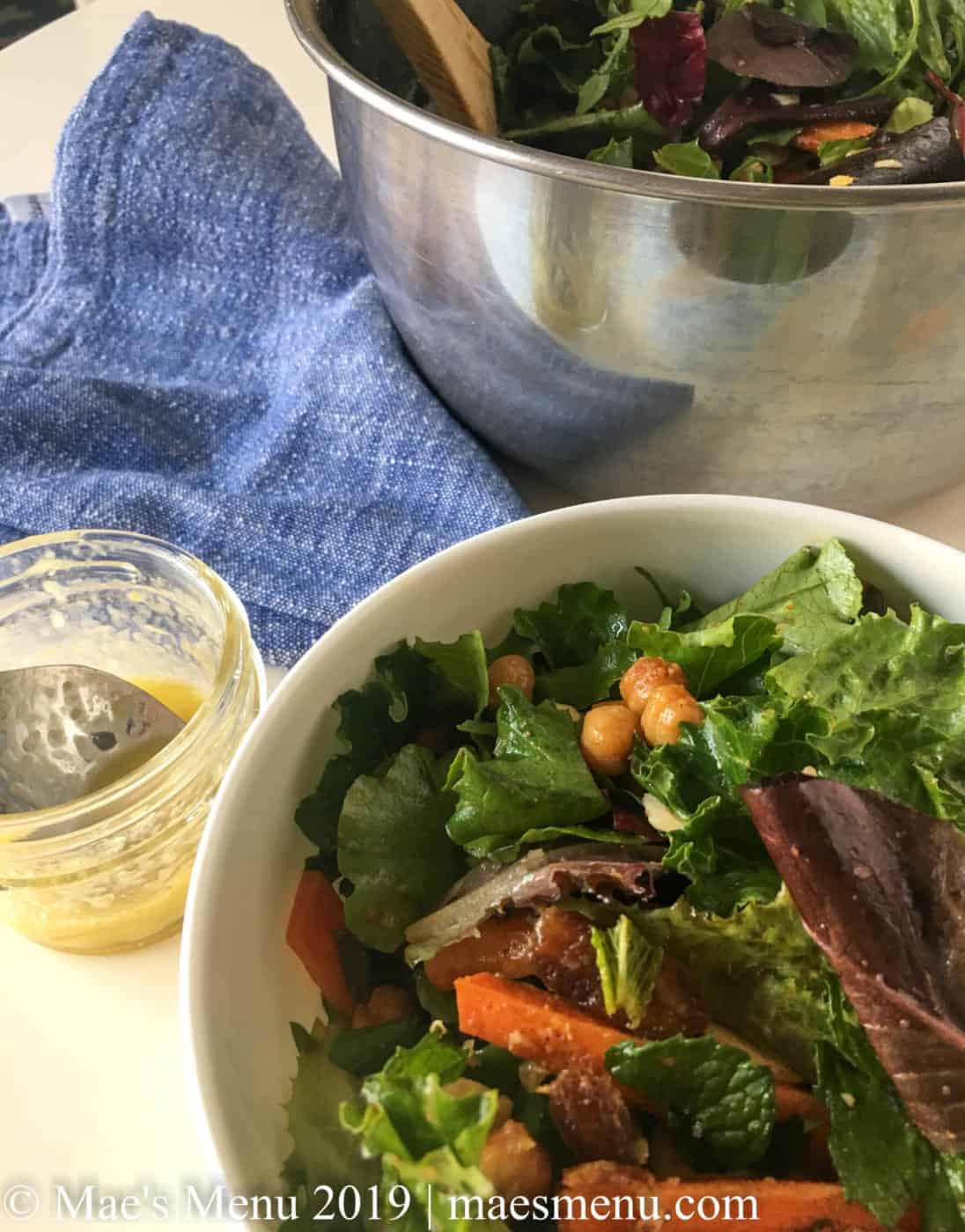 White salad bowl with green salad, chickpeas, and carrots in the foreground. Clear cup of vinaigrette and a spoon to the left of the bowl and a large metal bowl with the rest of salad behind that. Blue dishcloth in the back left of the photo.