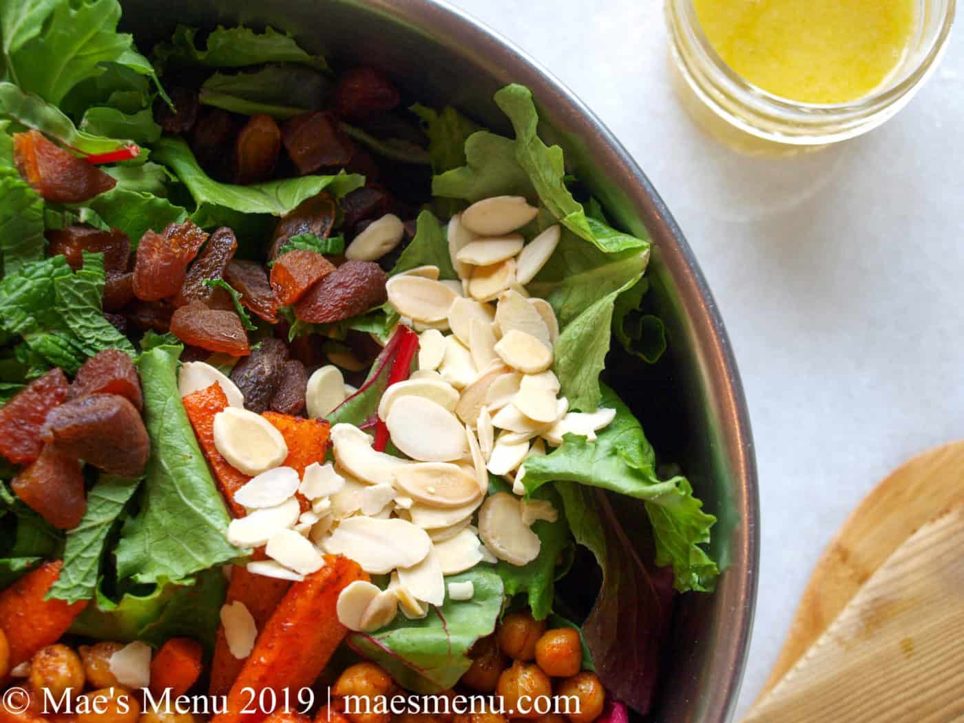 Metal bowl of green salad with chickpeas, carrots, almonds, and chopped dried apricots. Salad tongs and lemon dressing next to the bowl on the white counter.