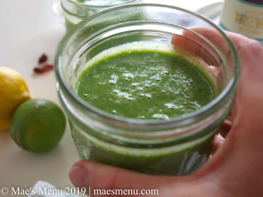 Hand holding a glass of Pineapple Green Smoothie. Fruit and other smoothies sit in the back.