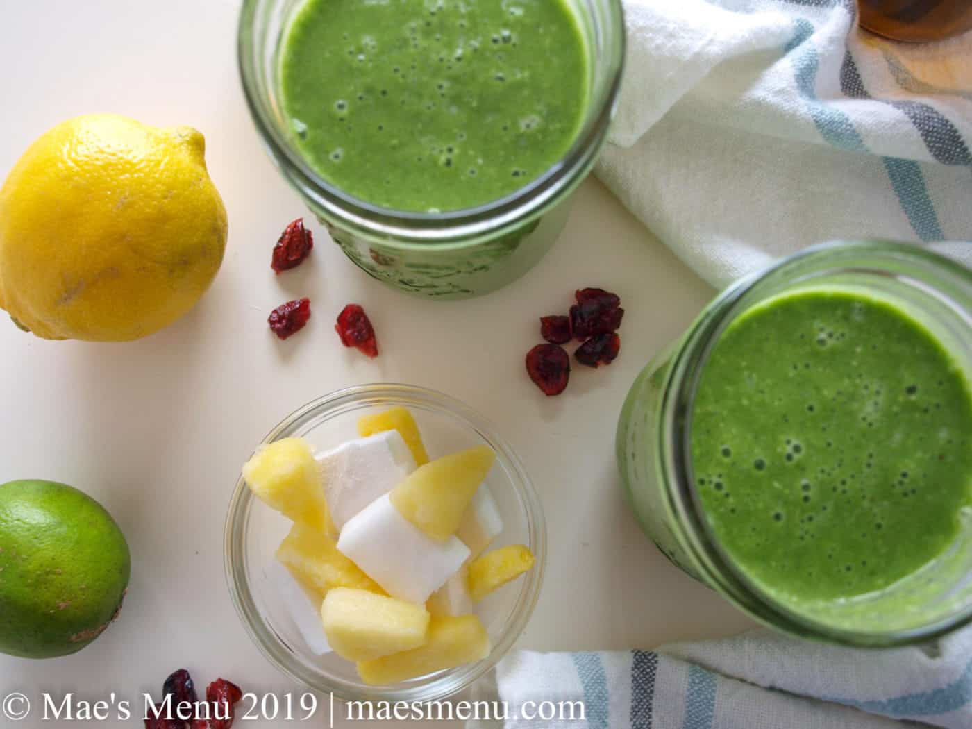 Two cups of Pineapple Green Smoothie sitting on a white table with cranberries, a blue and grey dish towel, a cup of frozen pineapple and coconut, and a lemon.