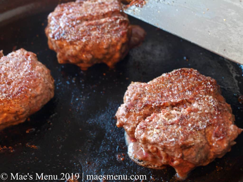 Three beef juicy hamburgers and a metal spatula sitting on a cast iron skillet.