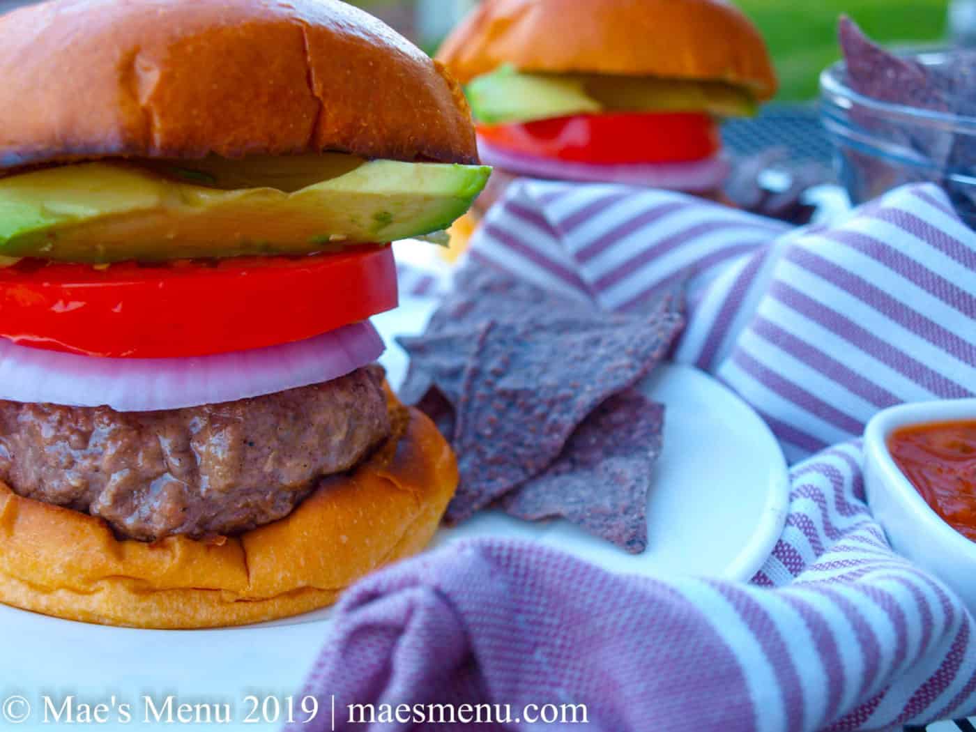Two tall burgers with blue corn tortilla chips and a purple striped dish towel.