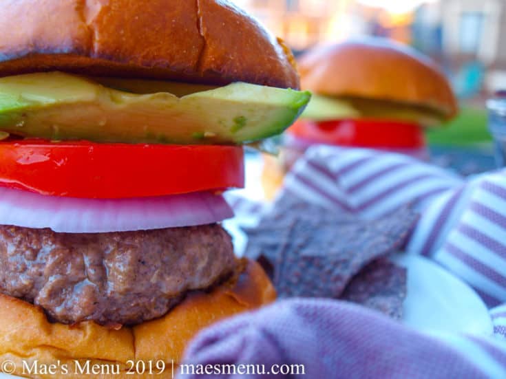 Two juicy hamburgers on white plates separated by a purple striped dish towel.