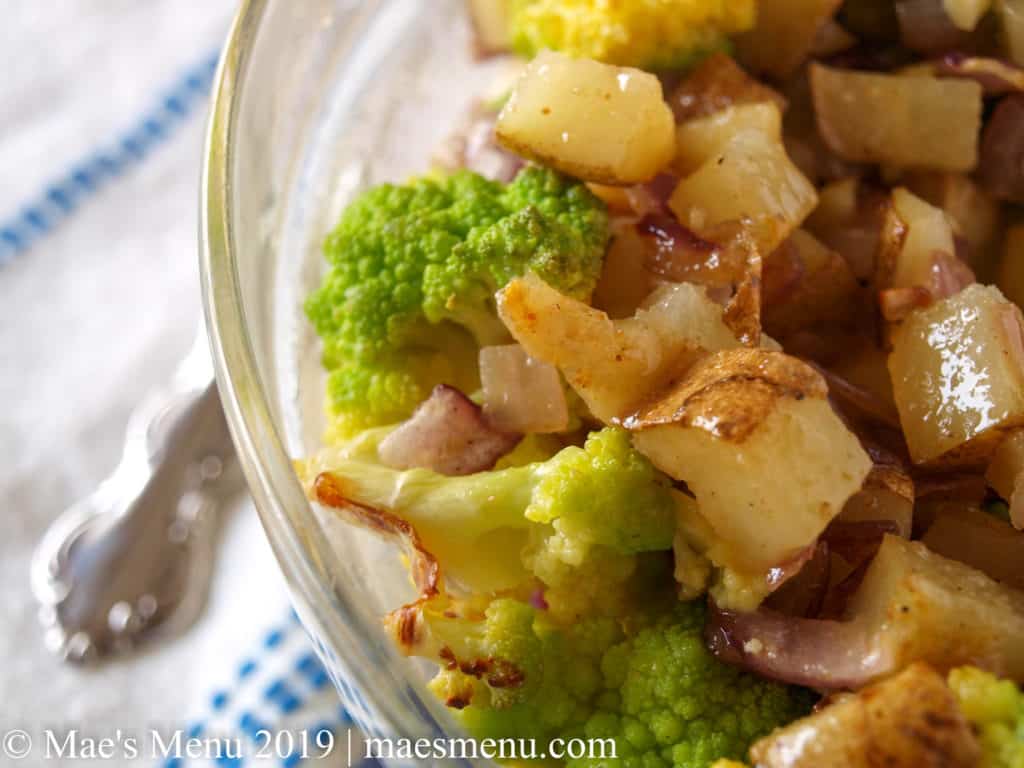 Clear bowl of roasted Romanesco Cauliflower and Potatoes tossed in a red wine vinaigrette. Blue and white dish towel with a spoon handle in the background.
