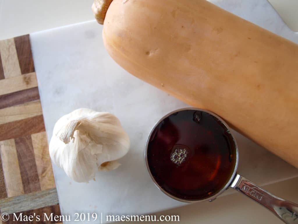 Butternut squash, a metal measuring cup of maple syrup, and a head of garlic on a marble and wood cutting board.