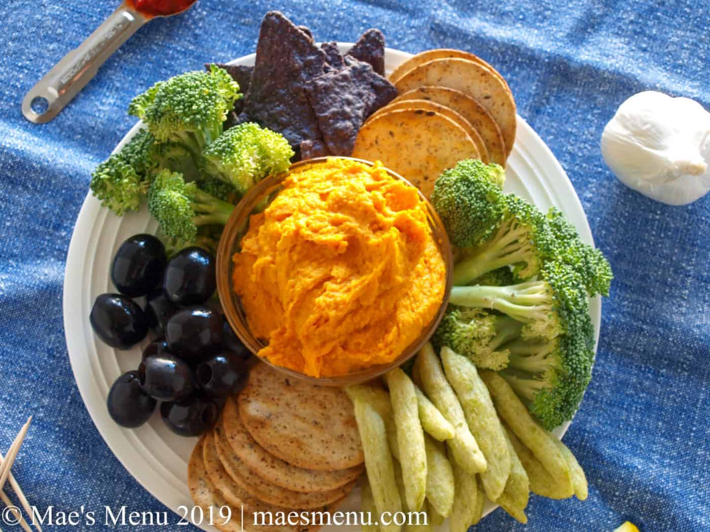 Sweet Potato Hummus with Harissa, veggies, and crackers on a white plate. Garlic clove, tooth picks, harissa, and lemons surround plate and sit on a blue dish towel.