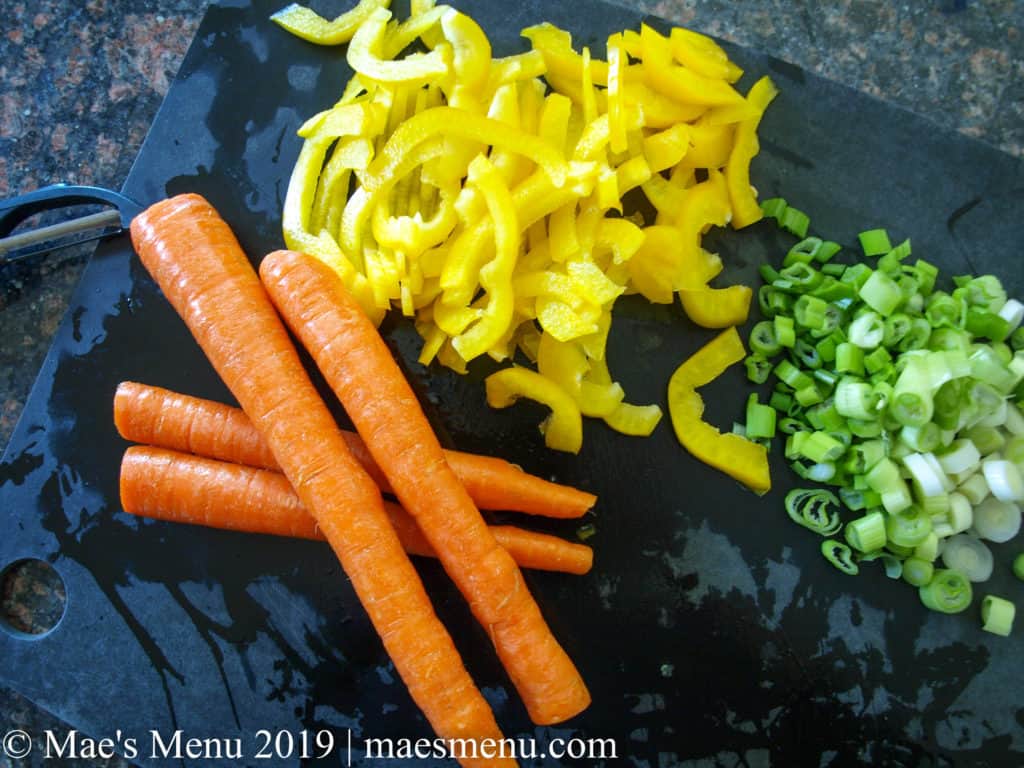 Sliced yellow peppers, green onions, and 4 carrots site on a black cutting board.