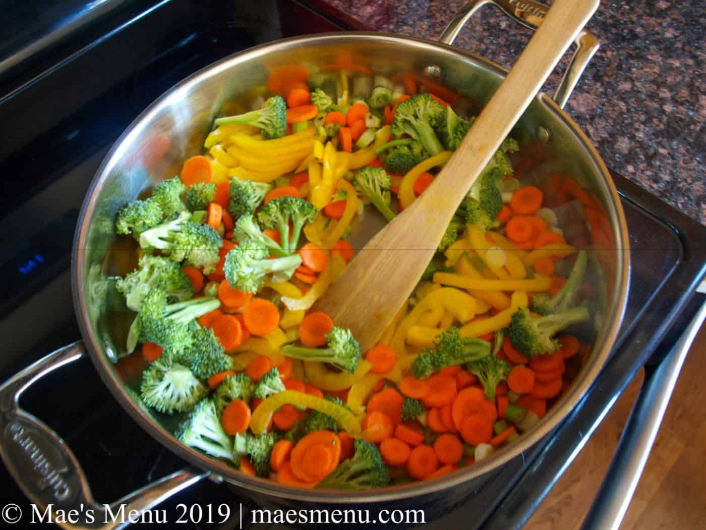 Broccoli, carrots, peppers, and green onions sauteing in a large metal pan.