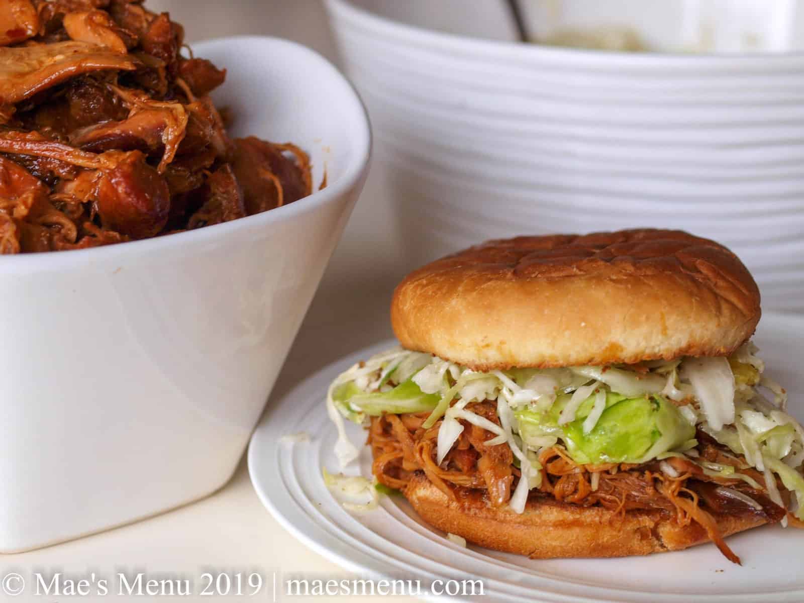 Honey BBQ shredded chicken sandwich on a white plate next to a bowl of bbq chicken.