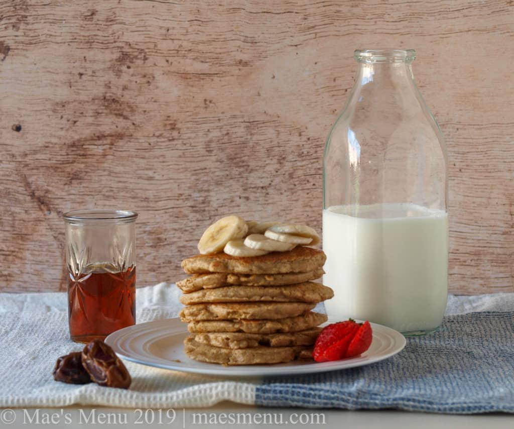 A plate with a stack of oatmeal blender pancakes. On top of the pancakes sits bananas and next to the pancakes sit strawberries. Next to the plate are dates, a cup of maple syrup, and a bottle of milk