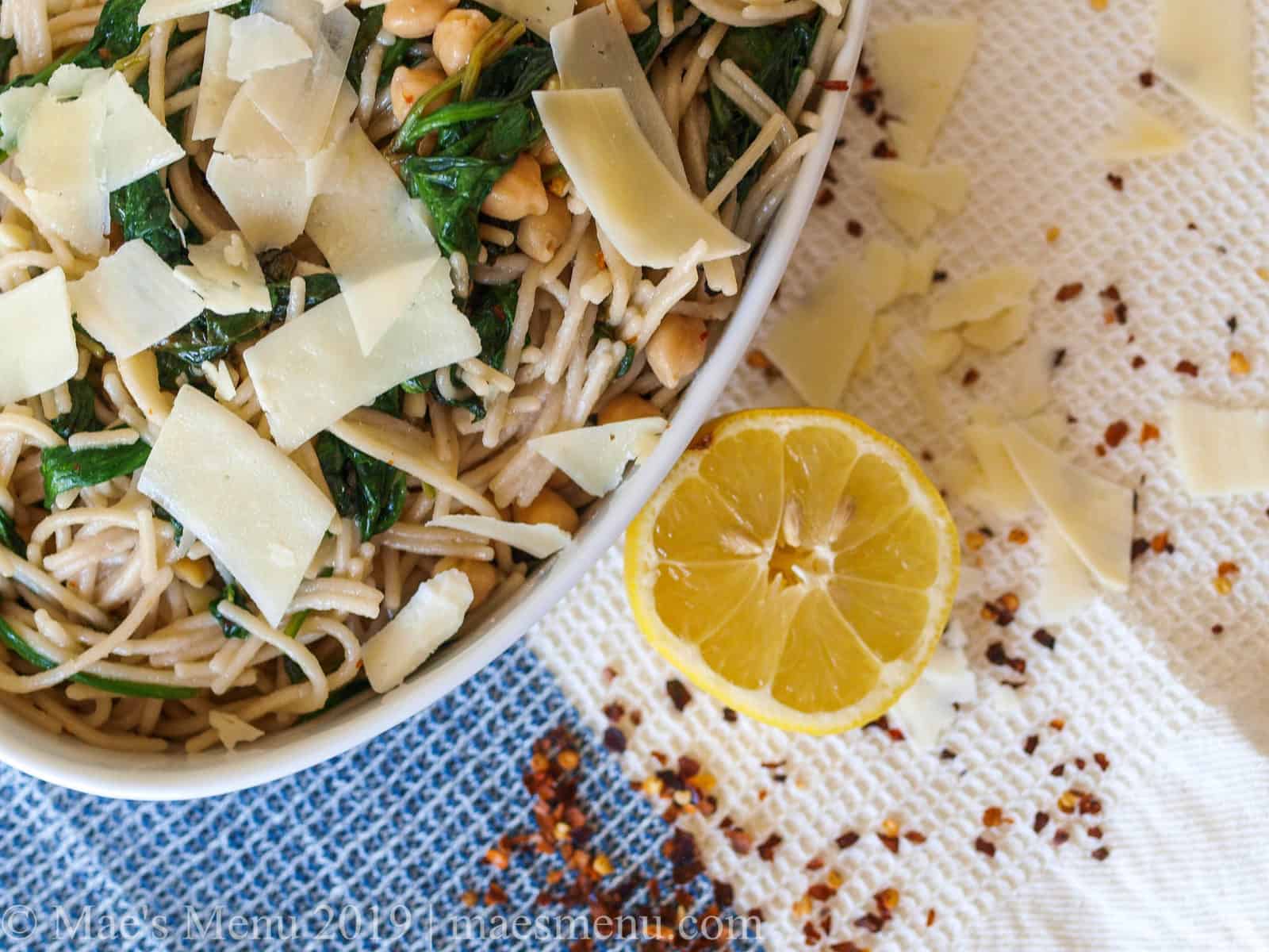 A white serving bowl of Spaghetti with Beans & Garlicky Greens next to a lemon, red pepper, and cheese.