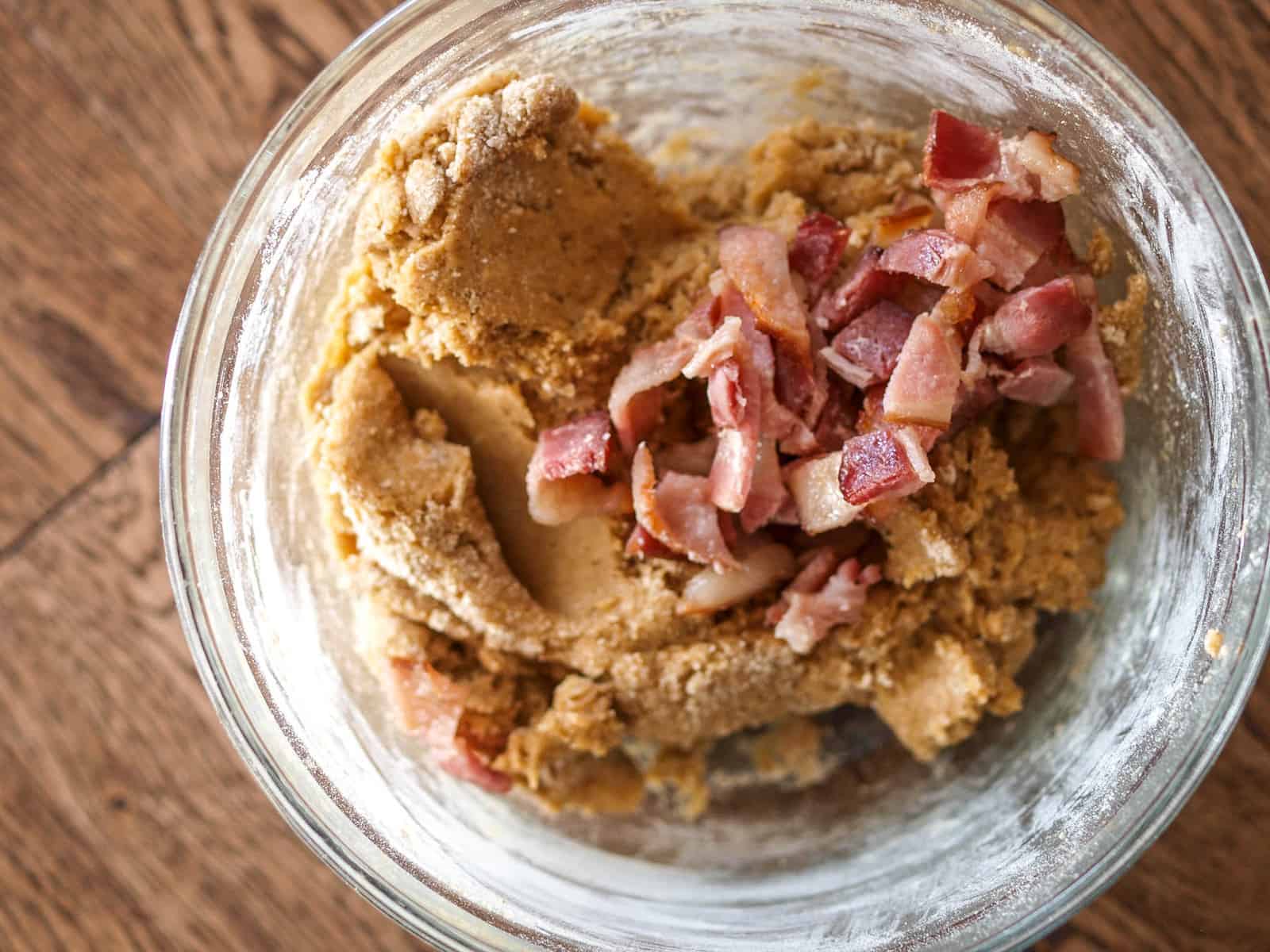 A large glass mixing bowl of peanut butter and bacon dog treat batter with chopped up bacon on top of it. 