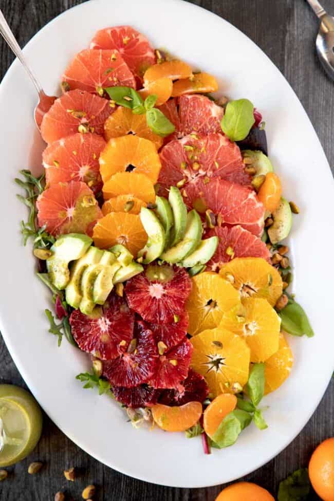 A large white platter of orange and grapefruit slices with avocado, pistachios, and baby basil leaves.