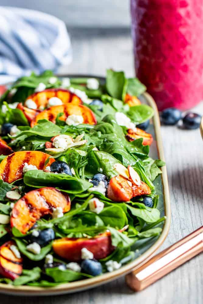 A large tan platter of spinach salad with grilled peaches, blueberries, and goat cheese. A glass container of blueberry dressing and a white and blue towel sits behind the salad. 