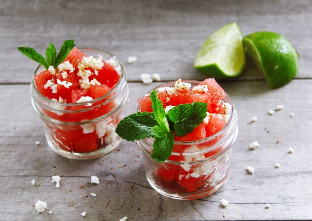 Two small glass cups of spicy watermelon salad next to two lime wedges.