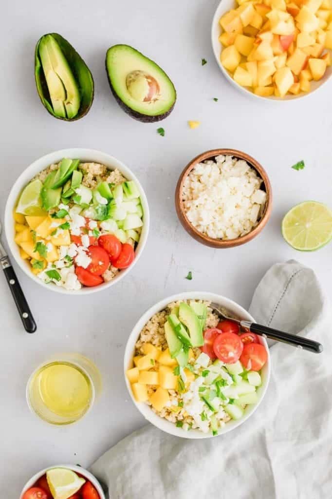 Bowls of Mango lime quinoa salad, a small container of feta cheese, a cup of chopped mango, a lime half, and avocado cut in half. 