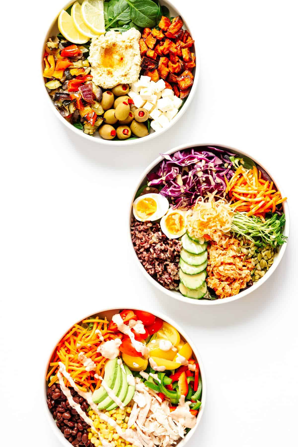 Three white bowls of nourish bowls with all different veggies, cheeses, and different ingredients in them.