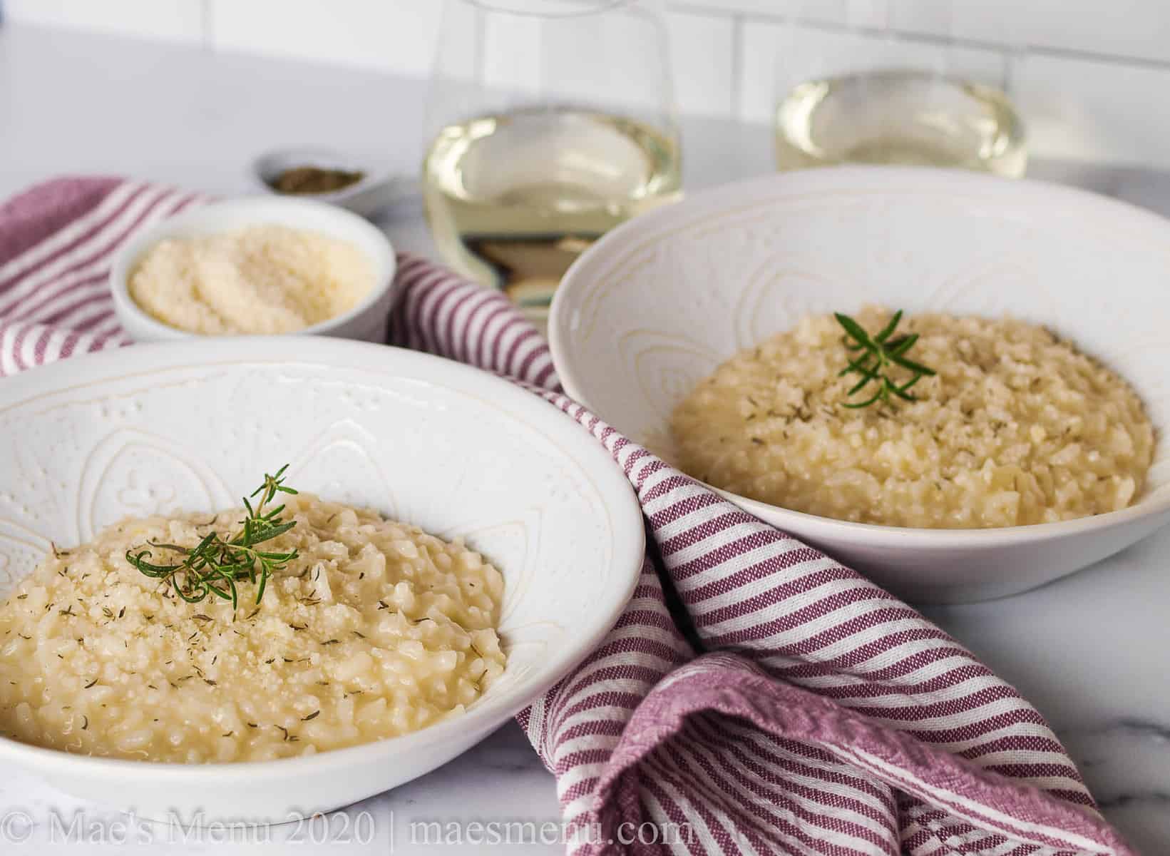 Two white bowls of risotto next to glasses of white wine, parmesan cheese, and a purple striped towel