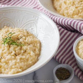 A large white bowl of risotto next to a cup of parmesan cheese and a spoonA large white bowl of risotto next to a cup of parmesan cheese and a spoonful of herbs.of herbs