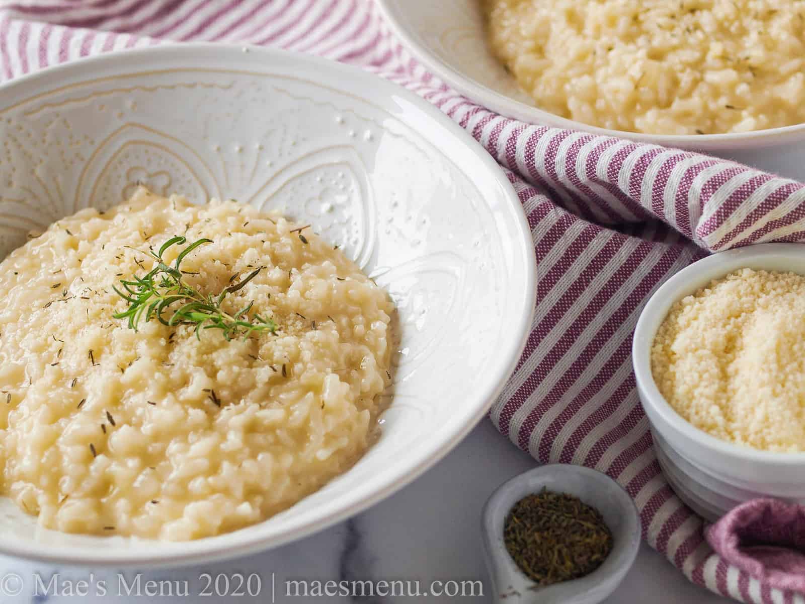 Two white bowls of risotto next to a cup of parmesan cheese and a spoonful of herbs