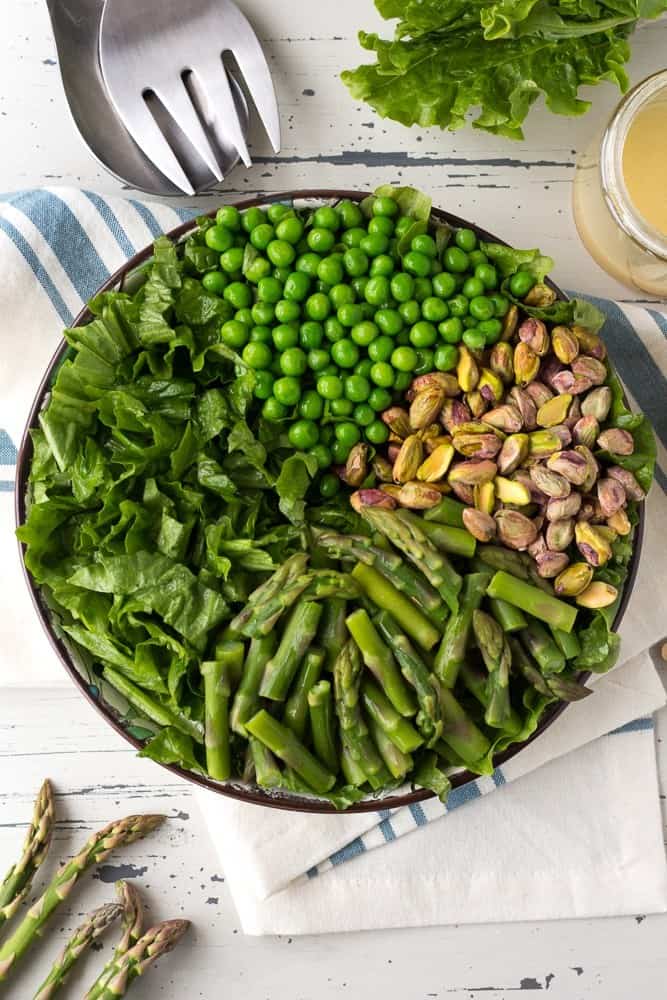A large bowl of greens covered with green peas, pistachios and asparagus. Next to the bowl sits salad tongs, a blue and white towel, more salad greens, and a cup of dressing.