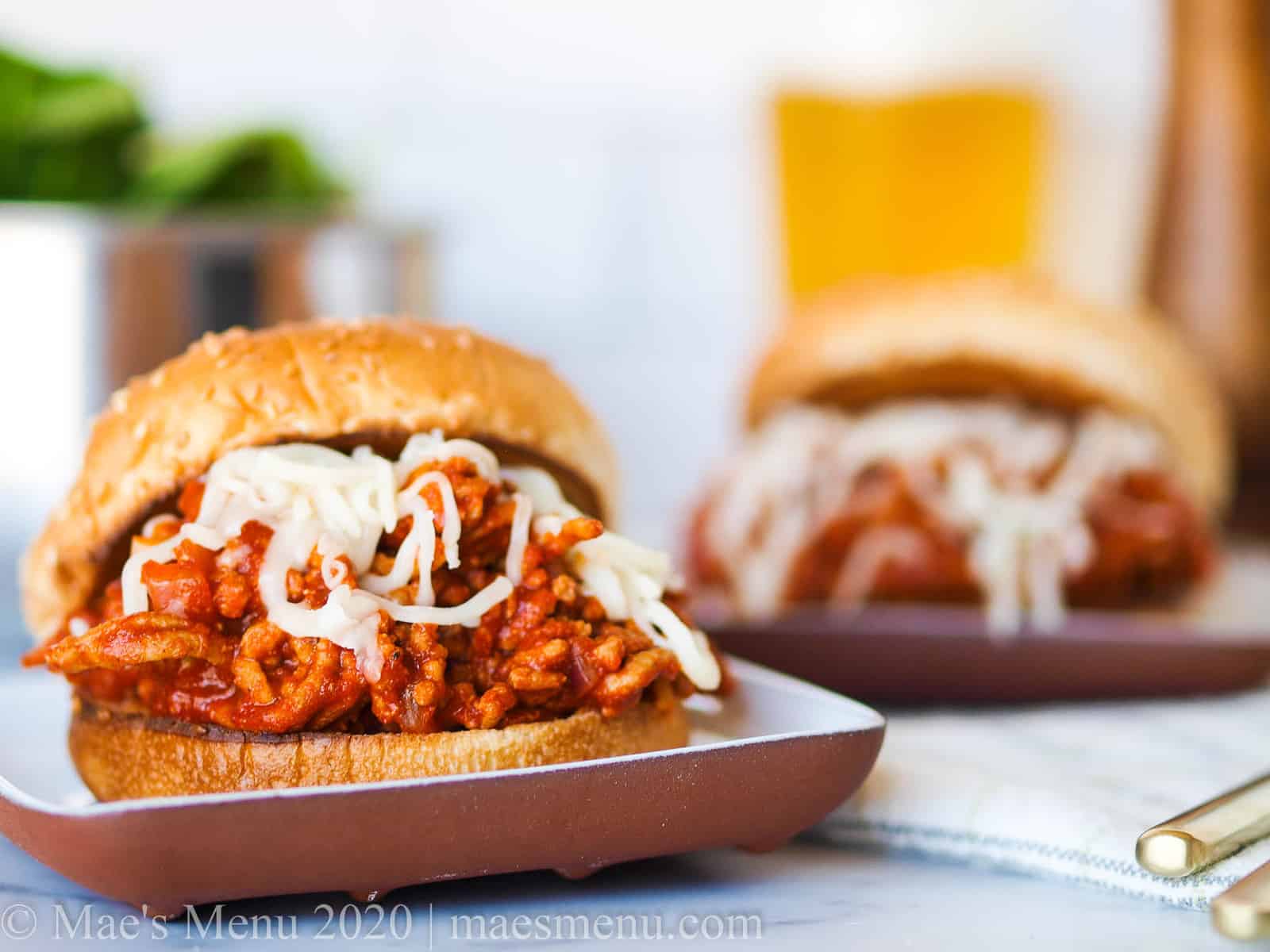 Two plates of sloppy joes on a dish towel with a pepper mill, a glass of beer, and a dish of salad in the background.