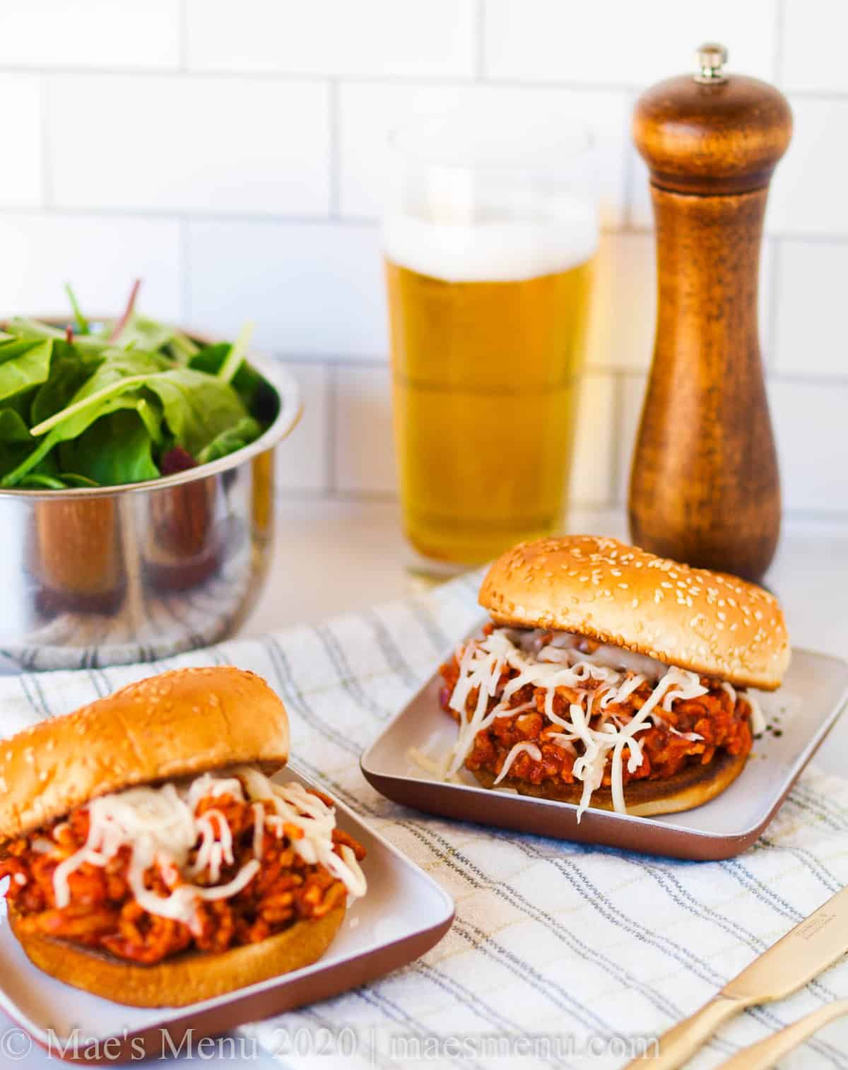 Two small plates of sloppy joes on a checkered napkin in front of a bowl of salad, a glass of beer, and a pepper grinder. 