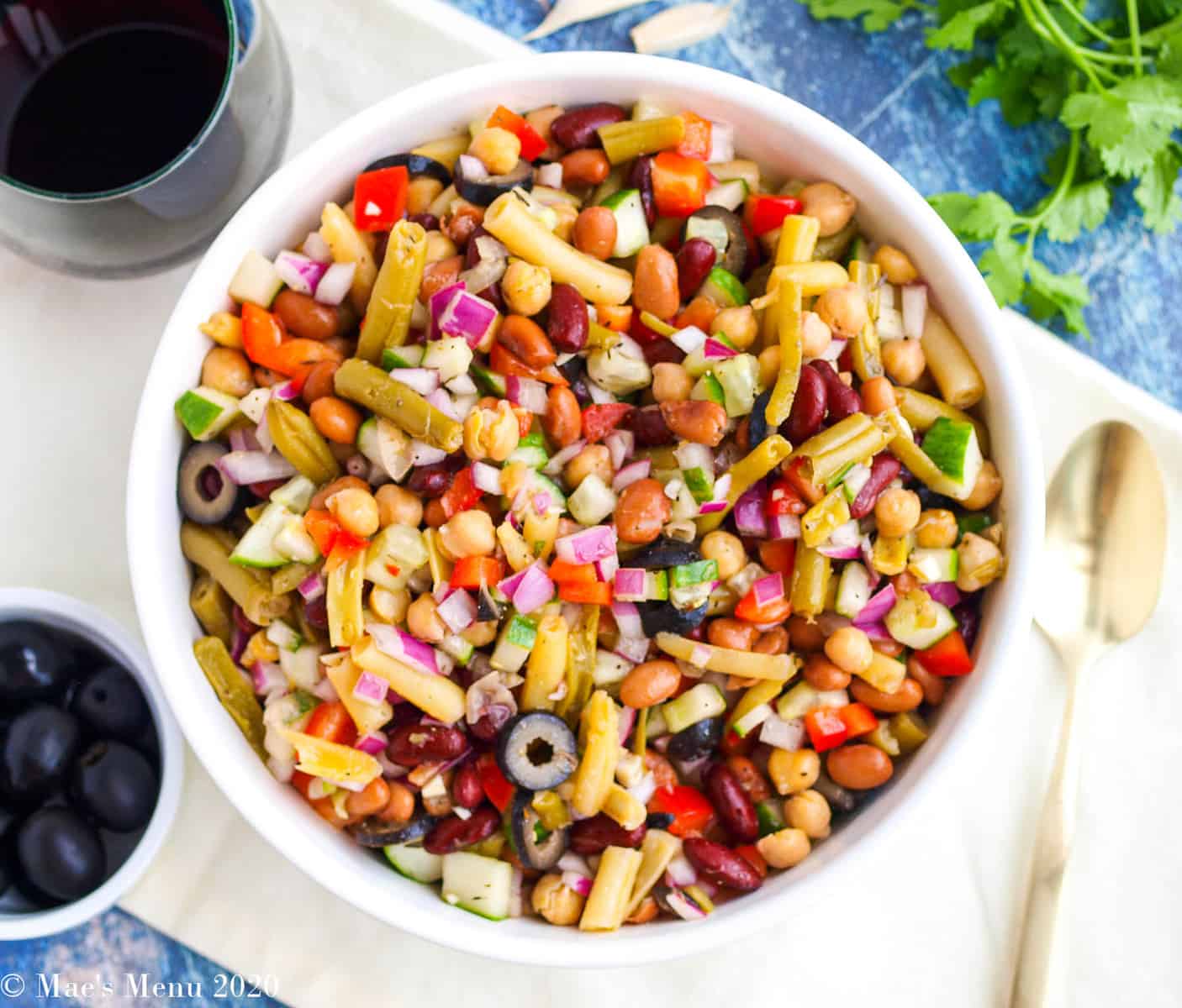 A large bowl of five bean salad with a dish of olives, a spoon, herbs, and a glass of wine next to it.