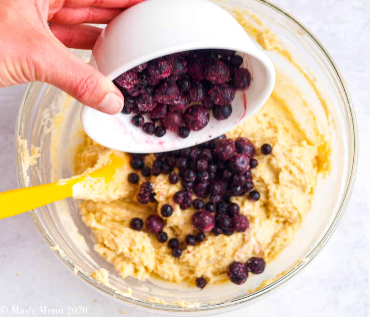 Pouring a small bowl of frozen blueberries into a large mixing bowl full of dough 