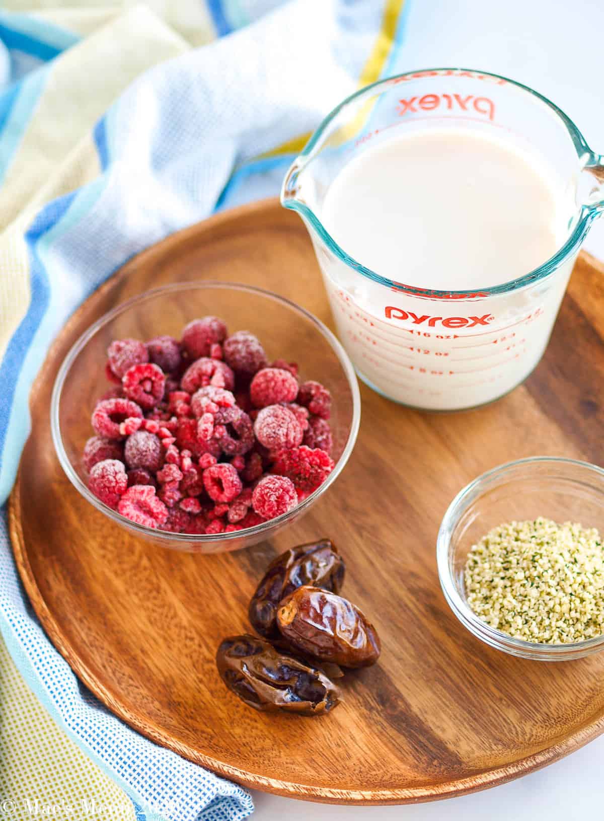 Some of the ingredients for a creamy raspberry smoothie: a cup of frozen raspberries, a 2 cup measuring cup of almond milk, 3 dates, and a small bowl of hemp seeds. 