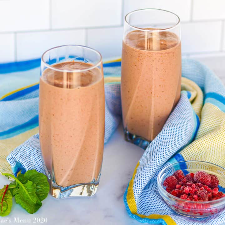 Two tall glasses of creamy raspberry smoothie next to a blue towel, a small cup of raspberries, and some baby greens