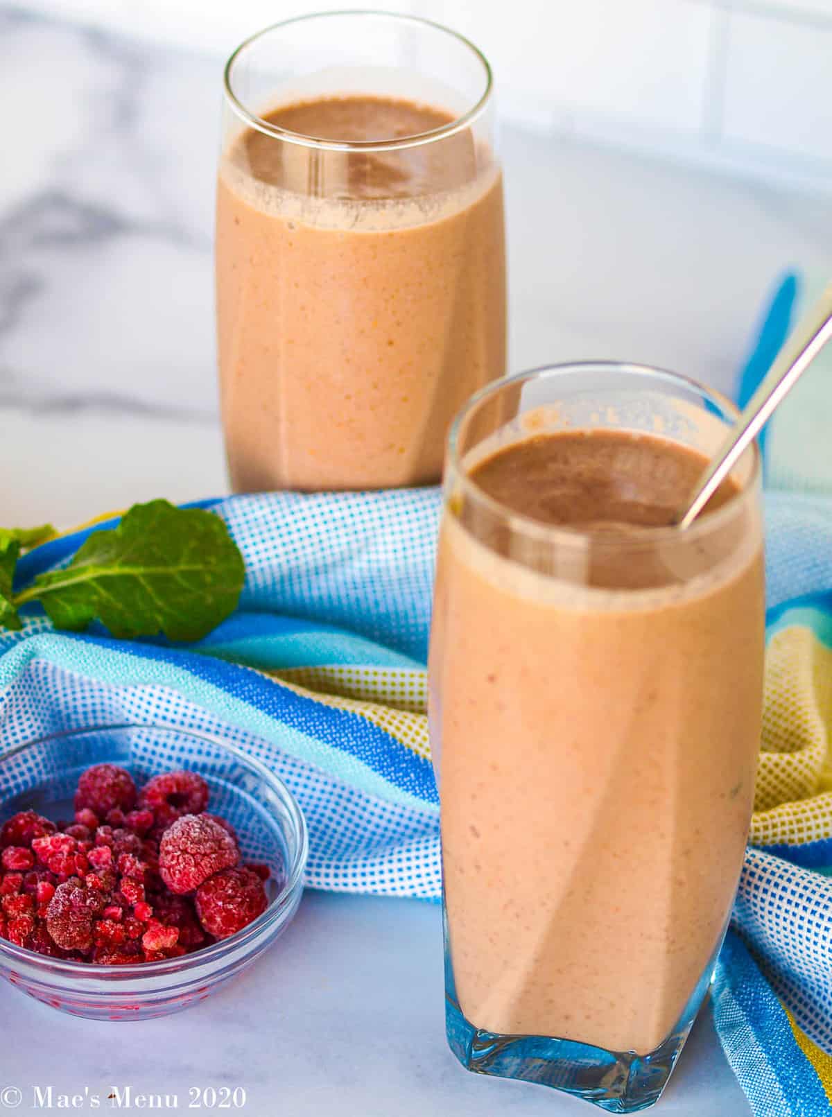 Two glasses of creamy raspberry smoothie with a blue dish towel between them. a Small dish of raspberries sits to the left of the glasses.