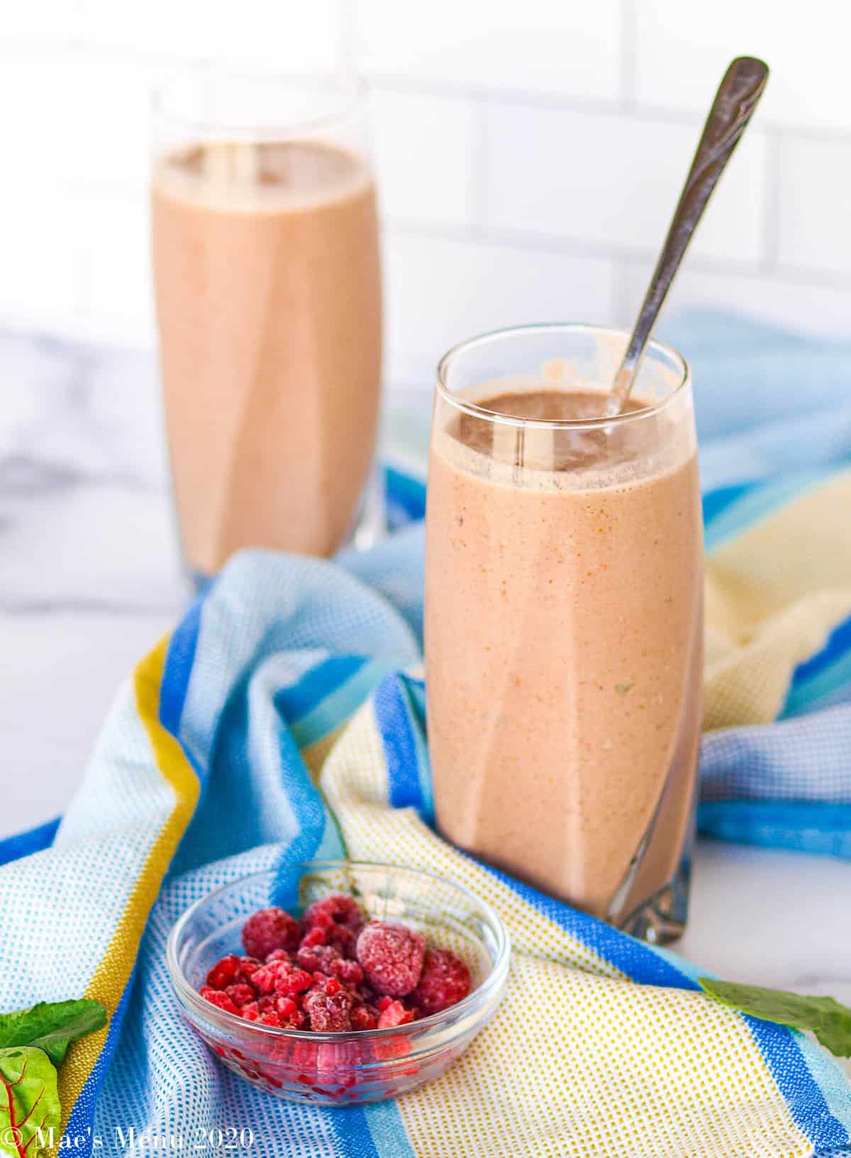 Two large glasses of creamy raspberry smoothie with a blue dish towel between them. In front of the glasses sits a small dish of frozen raspberries and a few baby greens.