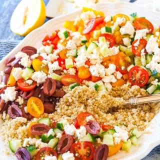A large white platter of Greek Quinoa Salad with a serving spoon in the salad. A lemon sits behind the plate.