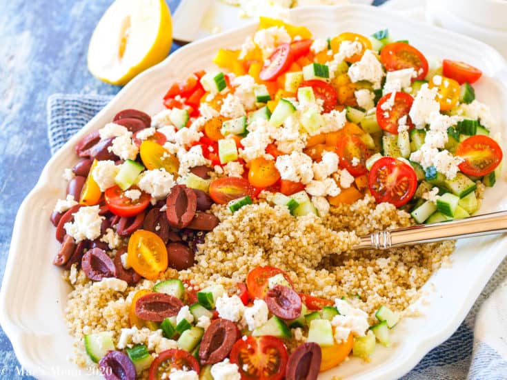 A large white platter of Greek Quinoa Salad with a serving spoon in the salad. A lemon sits behind the plate.