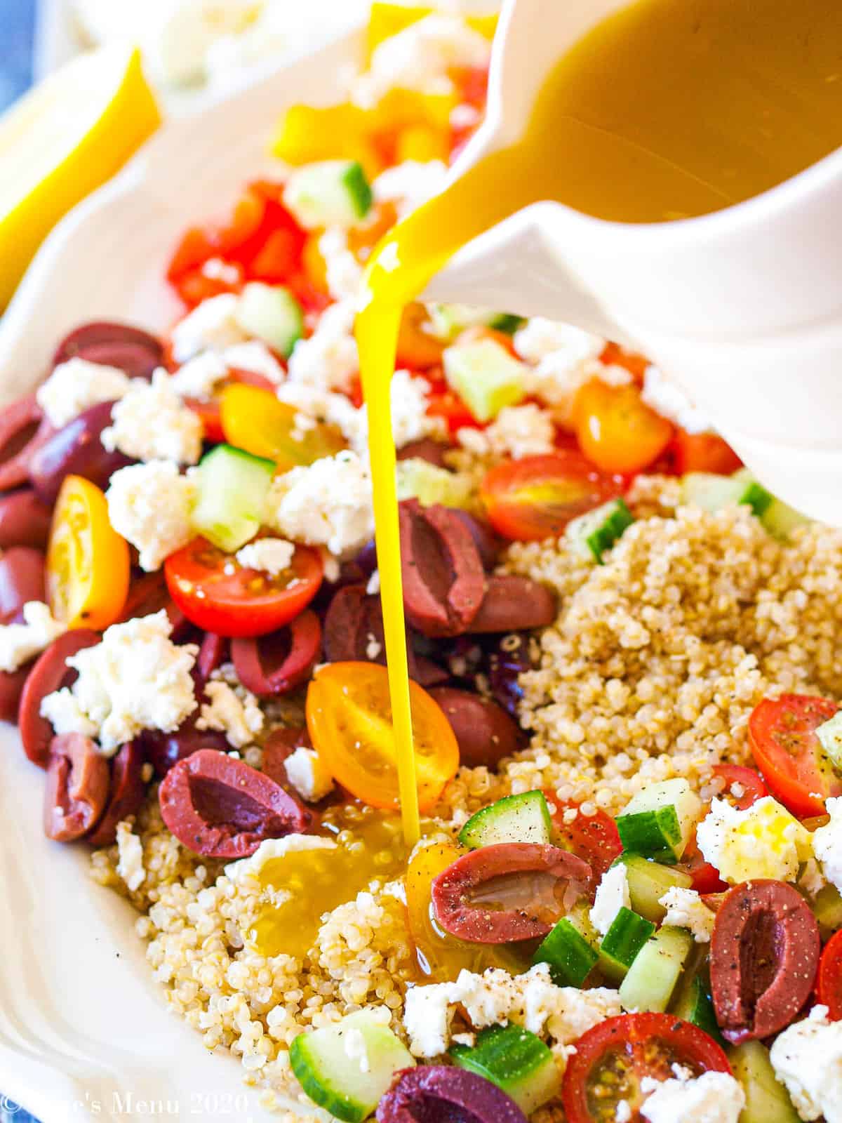 A platter of greek quinoa salad with a pitcher of dressing pouring over it.