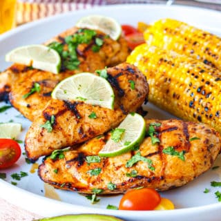 A large white platter with grilled chicken breasts covered in cilantro and limes next to grilled corn