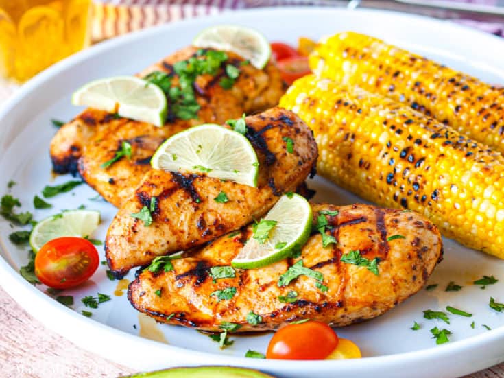 A large white platter with grilled chicken breasts covered in cilantro and limes next to grilled corn