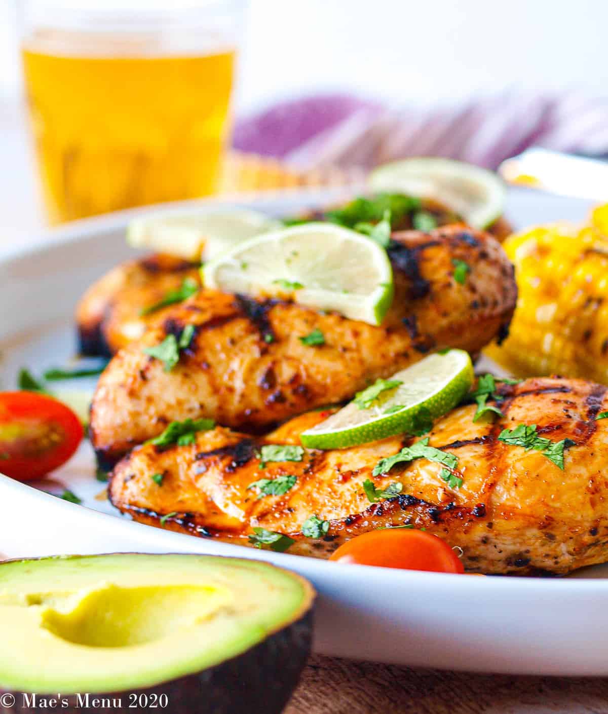 A side shot of a large white platter with grilled chicken breasts on it. In front of the platter sits a half of an avocado. On front of the chicken sits the lime slices and sliced cilantro. 