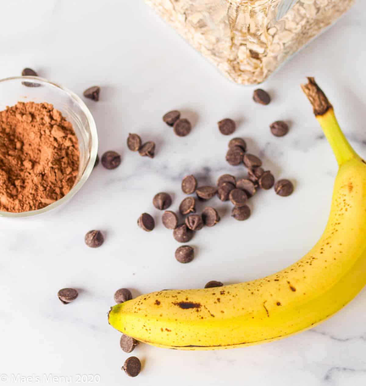 Chocolate chips, a banana, a small cup of cocoa powder, and the corn of a jar of oats on a granite counter. 