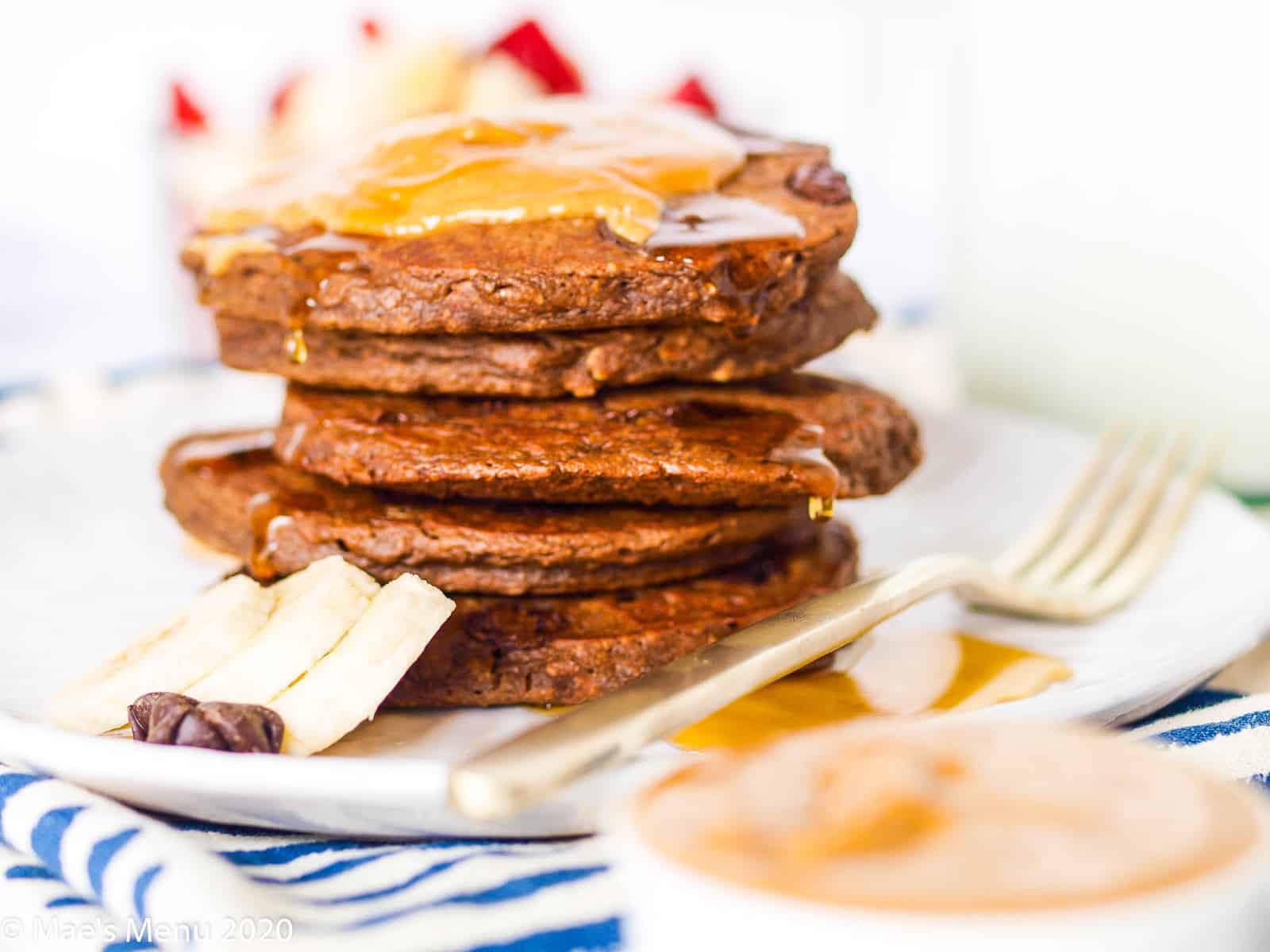 A stack of double chocolate chip banana pancakes with peanut butter and maple syrup on top. The pancakes sit on a white plate next to a fork.