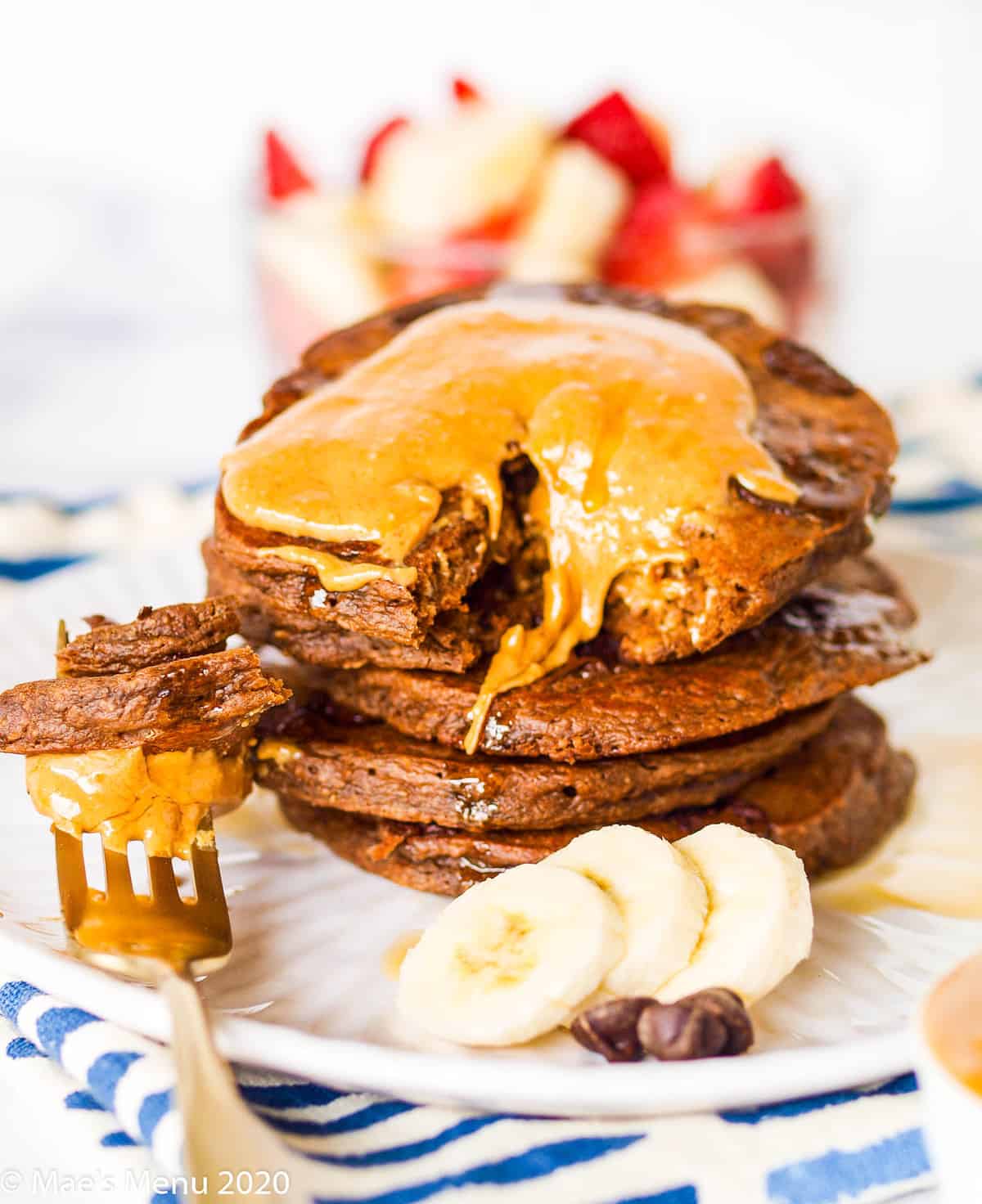 A plate of banana double chocolate pancakes covered in peanut butter. Beside the stack of pancakes sits a forkful of pancakes with peanut butter, banana slices, and chocolate chips